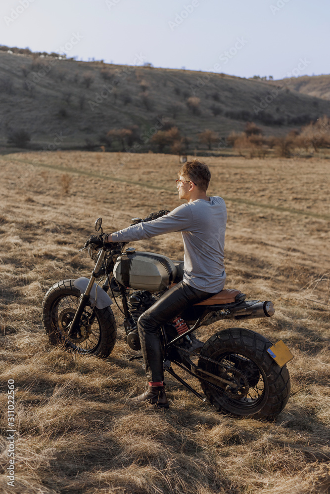 Back view of a motorcyclist sitting on a motorcycle in a countryside, admiring a landscape, in a sunset time. Vertical view. Back view of a motorcyclist sitting on a motorcycle in a countryside, admiring a landscape, in a sunset time. Vertical view.