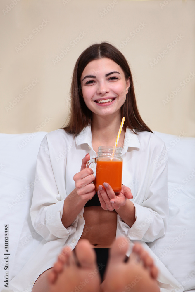 Beautiful girl drinking juice in bed and smiling. Breakfast concept at home or in the hotel. Copy space. Legs out of focus. Beautiful girl drinking juice in bed and smiling. Breakfast concept at home or in the hotel. Copy space. Legs out of focus.