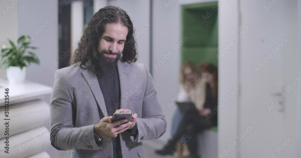 Portrait of handsome Middle Eastern man typing on smartphone keyboard, looking at camera and smiling. Confident office worker standing in open space. Business, success. Cinema 4k ProRes HQ. Portrait of handsome Middle Eastern man typing on smartphone keyboard, looking at camera and smiling. Confident office worker standing in open space. Business, success. Cinema 4k ProRes HQ.