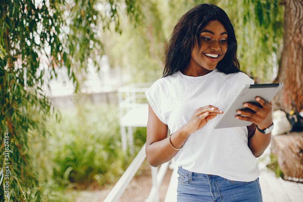Naklejka premium Cute black girl in a park. Lady in a white t-shirt and blue jeans. Woman with tablet