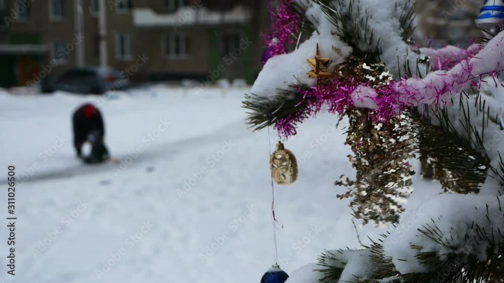 in the foreground a branch of a snowy spruce, in the background out of focus a grandmother and grandson ride a slide, slide riding, winter fun