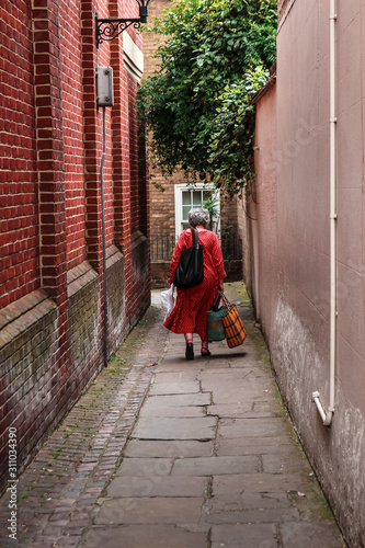 old woman in red dress with white polka dots walking with bags on a narrow street © Amaraz