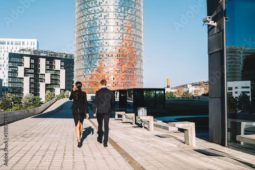 Coworkers against cityscape with high rise buildings Back view of confident diverse formal man and woman in suits walking on paved street against contemporary skyscraper in sunlight