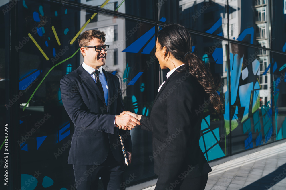 Plakat Happy contemporary coworkers shaking hands Smiling young man and diverse woman in black suits standing on city street in bright sunlight and shaking hands happily