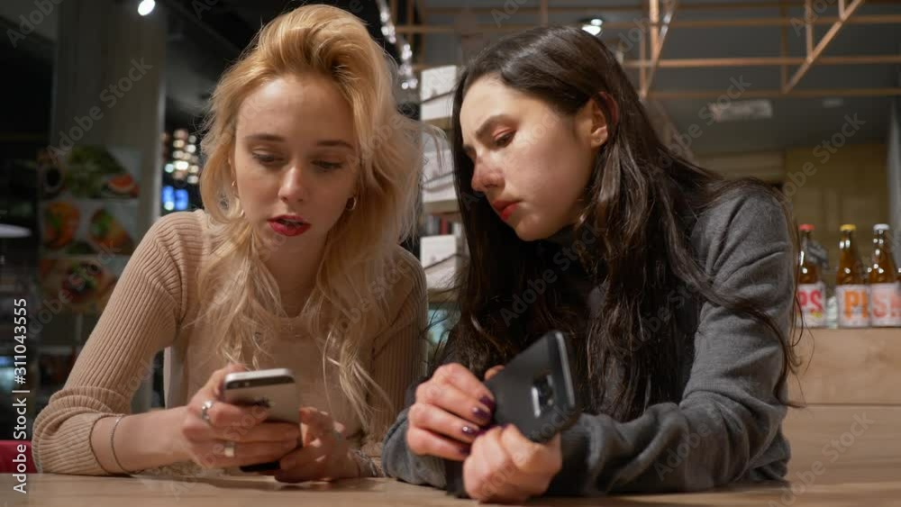 Two young pretty women girlfriends watch use mobile phones smartphones surfing internet social media while sitting in cafe waiting an order