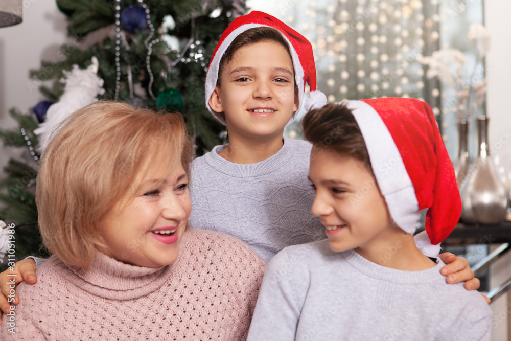 Cheerful little boy smiling to the camera hugging his twin brother and grandmother near Christmas tree Cheerful little boy smiling to the camera hugging his twin brother and grandmother near Christmas tree