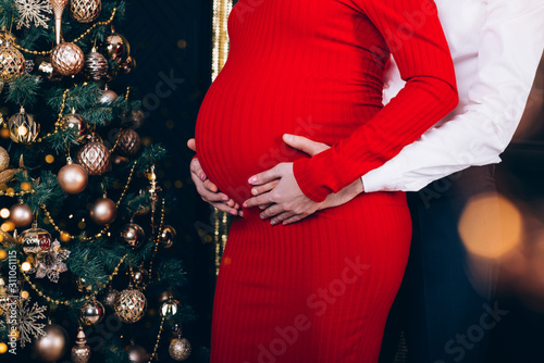 Parents hands touching pregnant belly near christmas tree decorated with golden toys. Cropped.