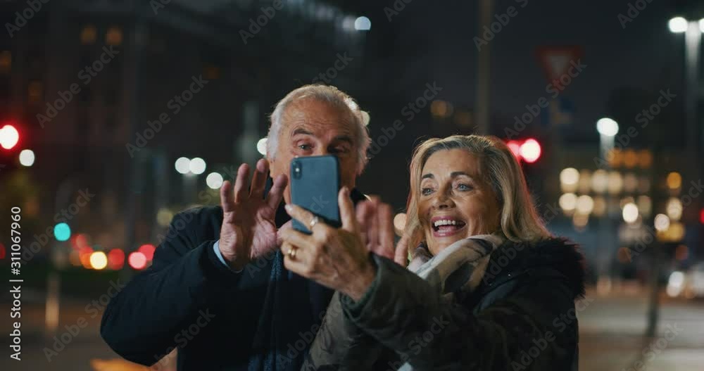 A happy senior couple is making a selfie or video call with a smartphone in a city center in the evening. A happy senior couple is making a selfie or video call with a smartphone in a city center in the evening.