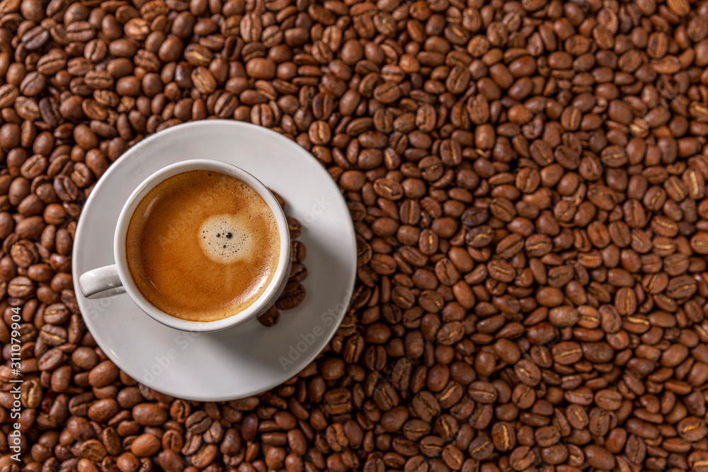 A small white cup of coffee stands on coffee beans. Fragrant pleasure. Background. Space for text. Top view. A small white cup of coffee stands on coffee beans. Fragrant pleasure. Background. Space for text. Top view.