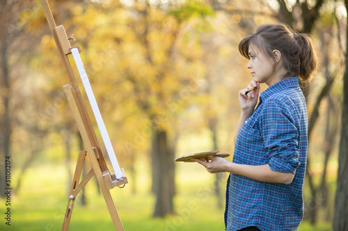 young woman artist drawing a picture on canvas on an easel in nature  a girl with a brush and a palette of paints working inspired by early autumn  a concept of art  hobby