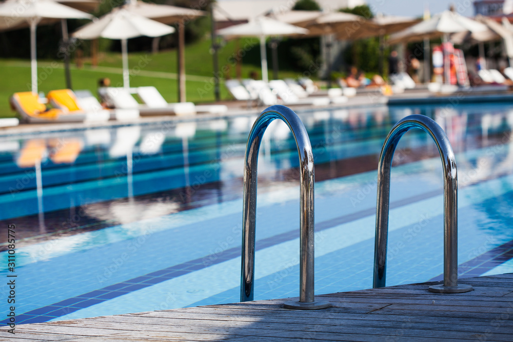 Obraz premium Outdoor large blue swimming pool on site with stairs to descend into the water. Sunbeds and sun umbrellas are visible in the background.