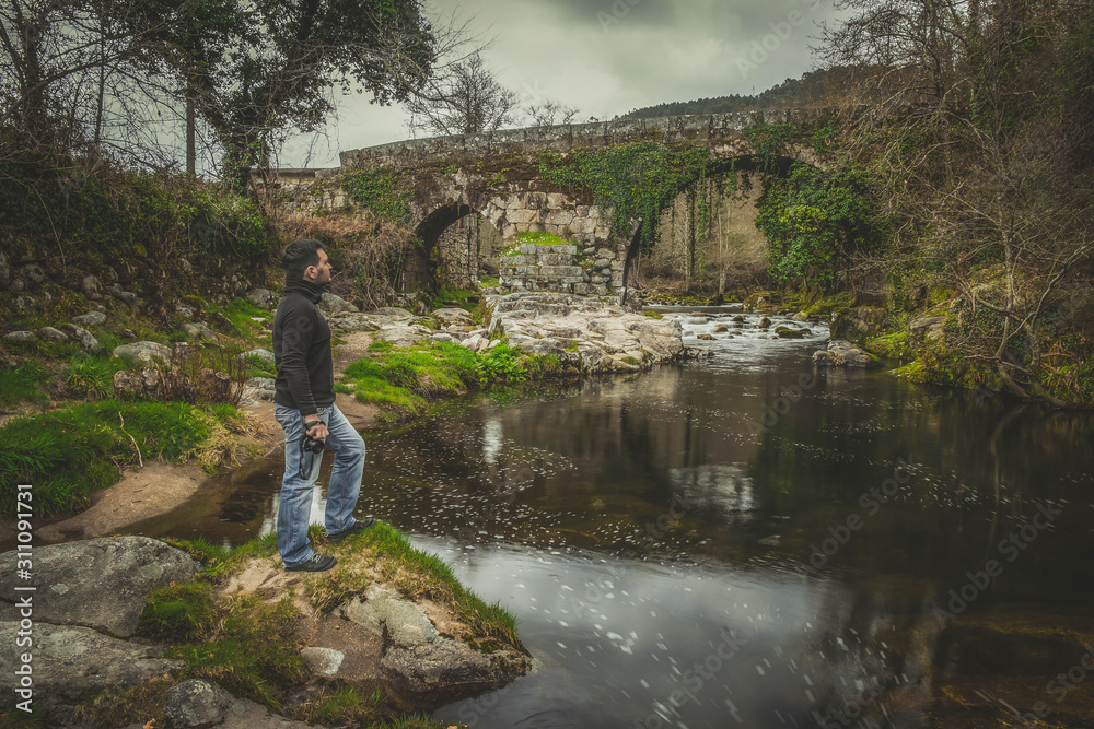 homem com mochila às costas a olhar para a paisagem ponte de pedra antiga