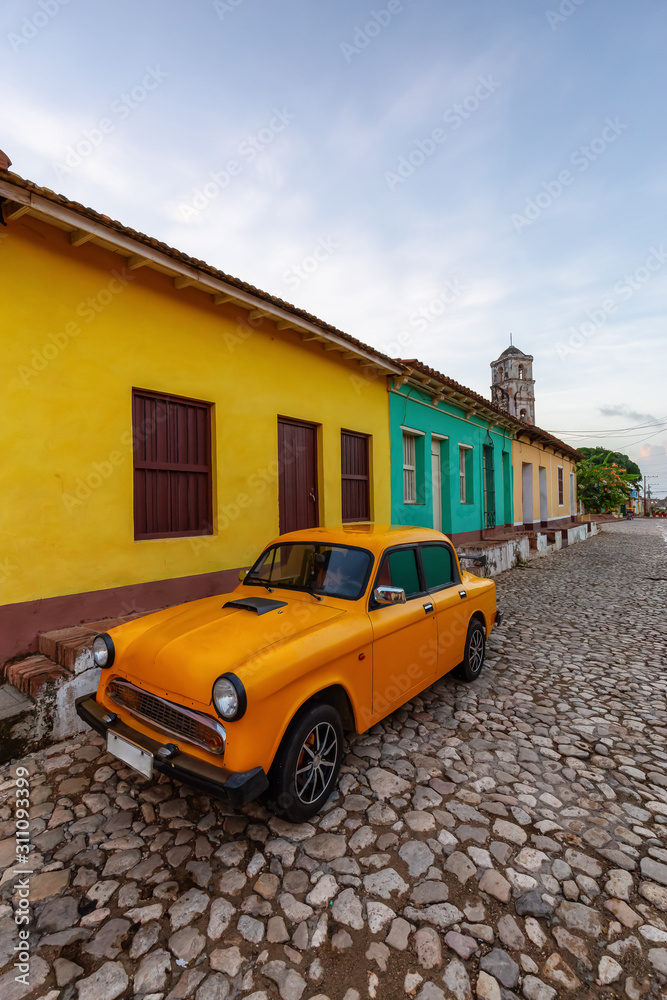 Fototapeta premium View of an Old Classic Taxi Car in the streets of a small Cuban Town with Church in the Background during a vibrant sunny sunrise. Trinidad, Cuba.