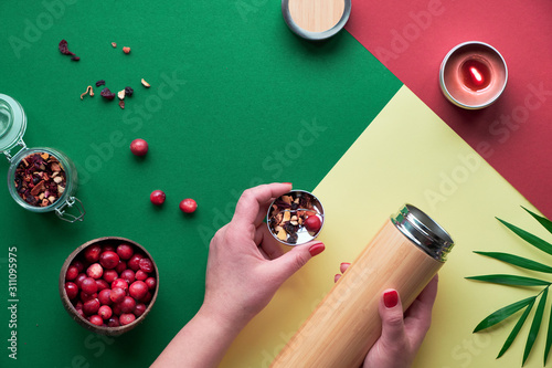 Zero waste tea in travel flask. Making herbal infusion in eco friendly insulated bamboo flask with herbal mixture and fresh cranberry berry. Trendy flat lay with hands, green red and yellow paper. photo