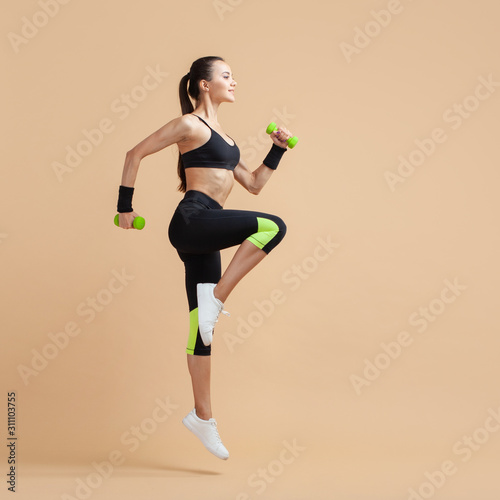 A young brunette woman is engaged in fitness, jumping with dumbbells, raising her knees high, on a peach background.