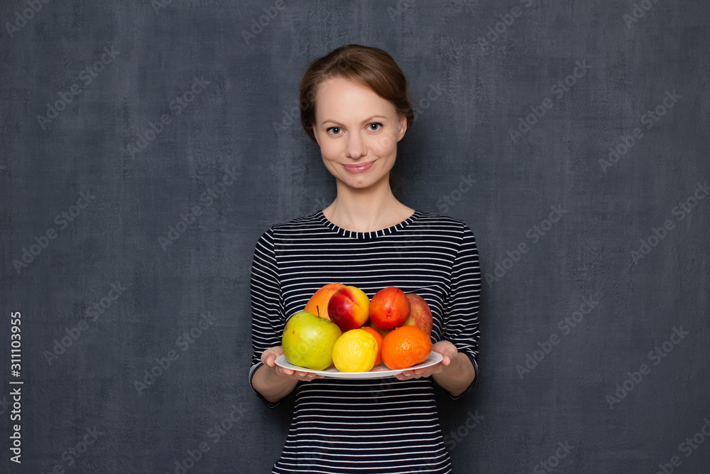 Portrait of happy young woman smiling and holding plate of ripe fruits
