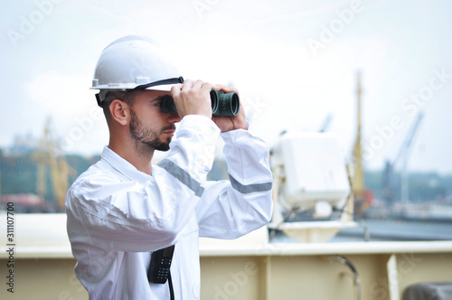 The navigator, the third assistant to the captain, is looking through binoculars in a white overalls and white helmet on the ship.