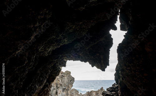 Cave in volcanic rock in Porto Moniz, a tiny resort village in Madeira. Arrow or church like a keyhole in the cliff that shows the coastline and rough sea water. Dark frame around autumn day on ocean.