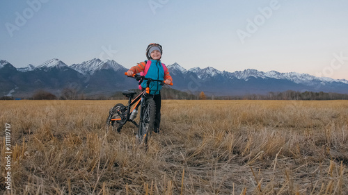 One caucasian children walk with bike in wheat field. Little girl walking black orange cycle on background of beautiful snowy mountains. Biker stand with backpack and helmet. Mountain bike hardtail.