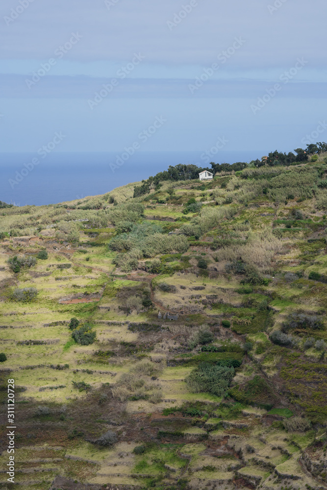 Aerial view onto tiny flat coastal area between steep vertical cliff and Atlantic Ocean. Tiny crop fields and cottages of local farmers far below on the shore washed by waves. Achadas Da Cruz, Madeira