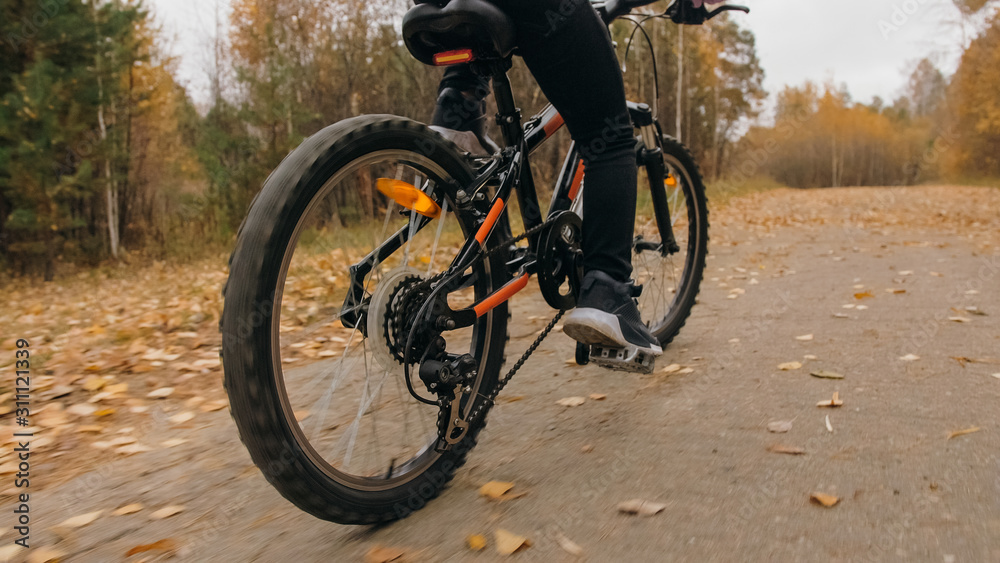 Fototapeta premium One caucasian children rides bike road in autumn park. Little girl riding black orange cycle in forest. Kid goes do bicycle sports. Biker motion ride with backpack and helmet. Mountain bike hardtail.