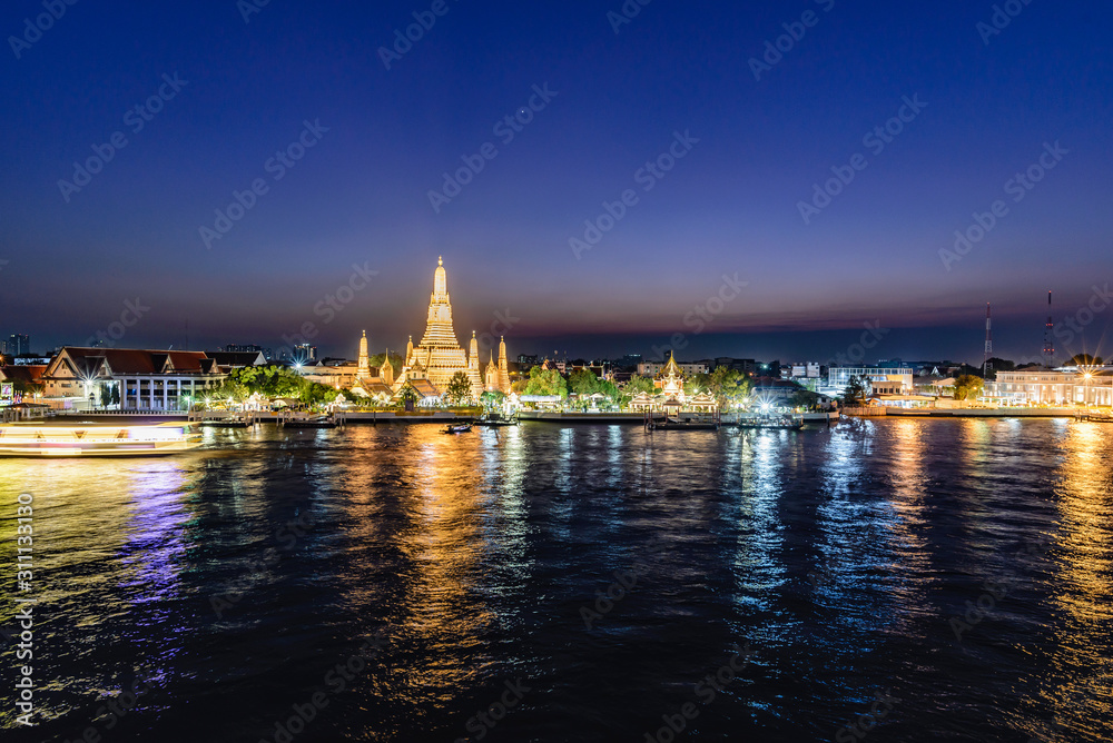 Night atmosphere of Wat Arun by the Chao Phraya river with golden light emitting a beautiful glow. There are tourists rushing through the front.