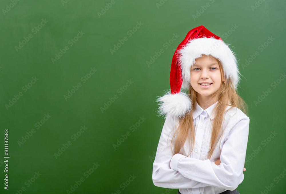 Smiling girl wearing a red christmas hat stands near green chalkboard. Empty space for text Smiling girl wearing a red christmas hat stands near green chalkboard. Empty space for text