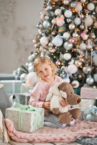 The girl sits near the Christmas tree and hugs a wolf doll in the skin of a s
