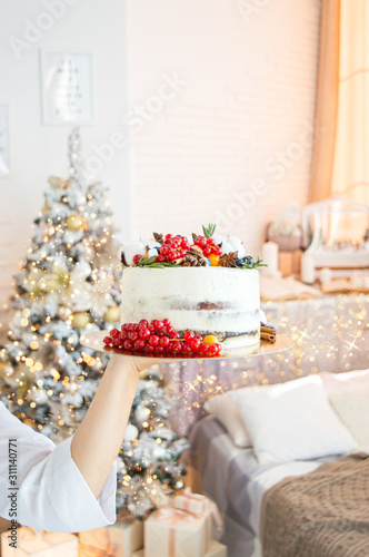 Cake decoration with lingonberry berries, blueberries and cotton. Women's hands in a frame in a bright kitchen. Christmas treat for the holiday. Lights of garlands.