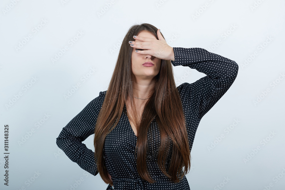 Charming caucasian woman covering eyes with hand, keeping hand on waist over isolated gray background wearing casual black dress with white polka dot. People lifestyle concepte. Charming caucasian woman covering eyes with hand, keeping hand on waist over isolated gray background wearing casual black dress with white polka dot. People lifestyle concepte.