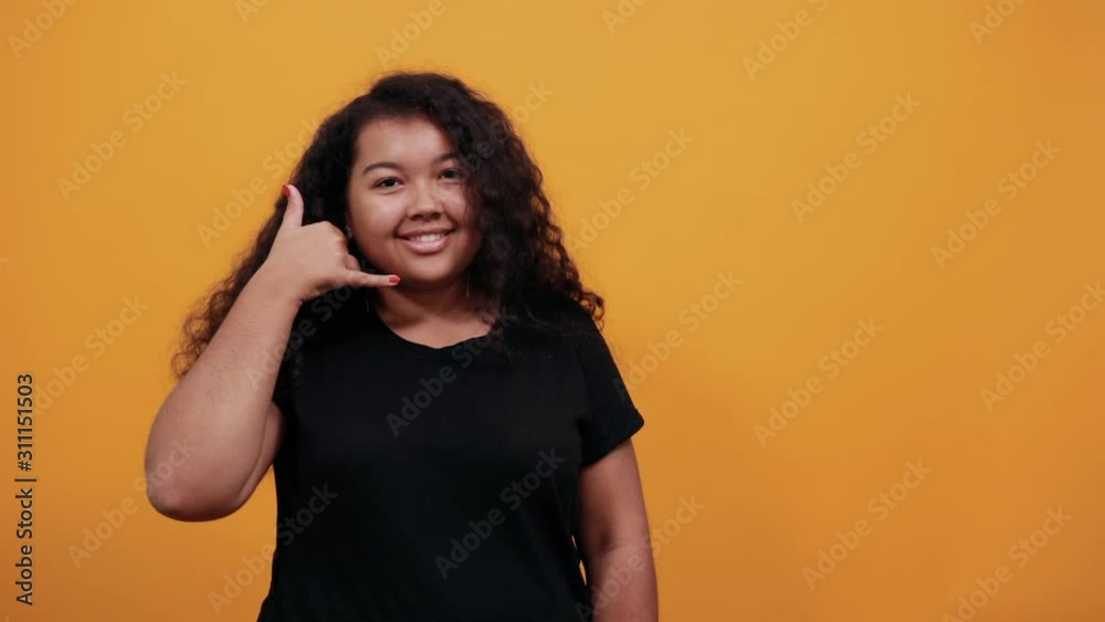 Afro-american young lady with overweight over isolated orange background wearing fashion black shirt doing telephone gesture near face, looking at camera. People lifestyle concept. Afro-american young lady with overweight over isolated orange background wearing fashion black shirt doing telephone gesture near face, looking at camera. People lifestyle concept.