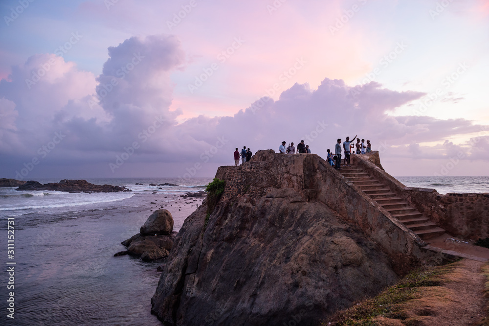 The tourists stand on the wall of Galle Fort at the ocean's coast and watch the sunset, Sri Lanka.