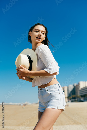 putting her ass back, the girl jokingly looks at her while holding a hat near her chest against the background of sand and high-rise buildings