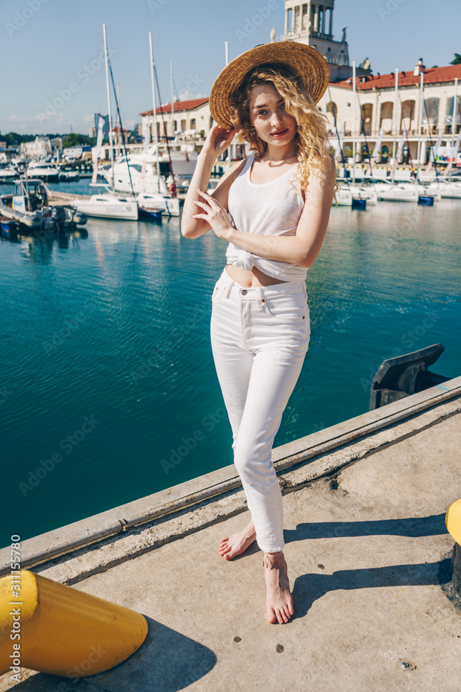 a girl in white trousers and a T-shirt stands barefoot on a pier against the background of the sea terminal