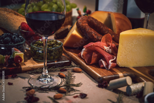Table with gourmet ingredients, glass of wine, variety of cheeses, cold meats, grapes, raspberry, blackberry and artisan bread on a rustic background