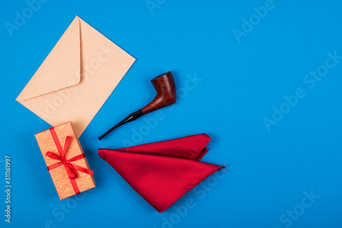 Gift box with bow, red pocket square, smoking tobacco pipe and craft paper envelope on blue background.