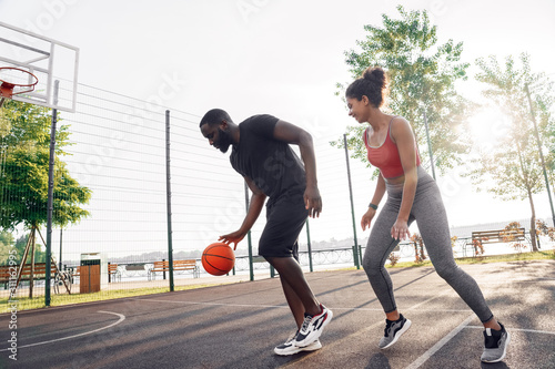 Outdoors Activity. African couple guy dribbling while girl defencing backdoor happy on basketball court in sunlight