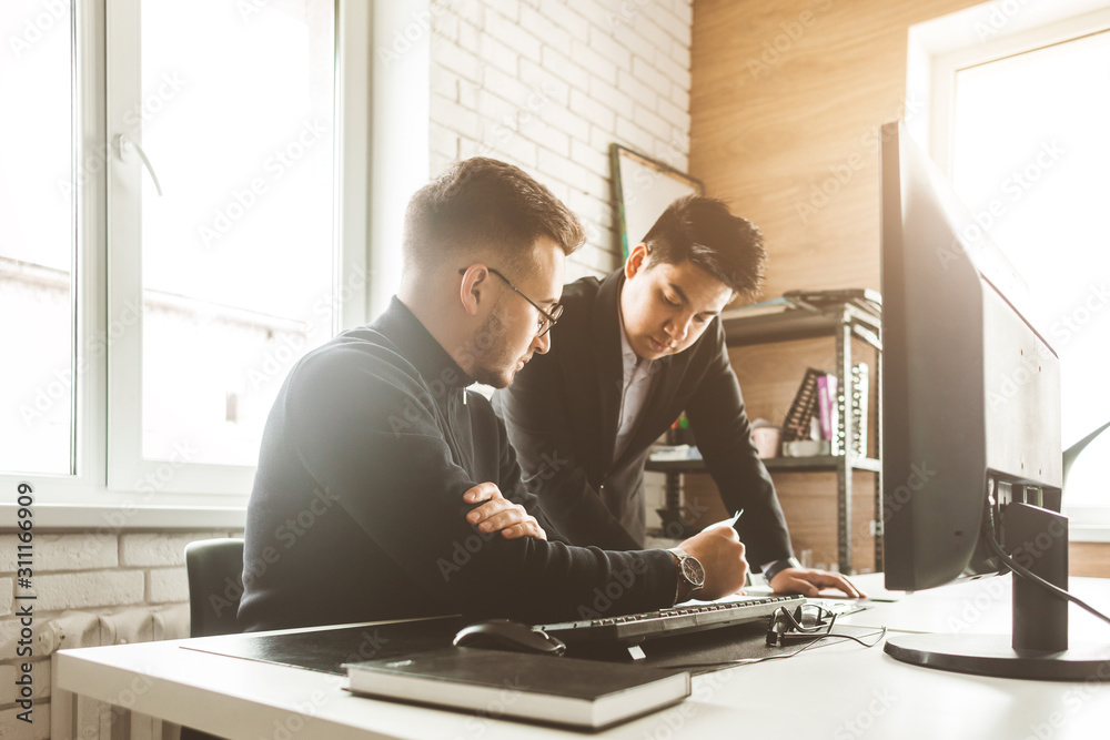 Young office worker sitting at desk, using computer. Two business man talking Young office worker sitting at desk, using computer. Two business man talking