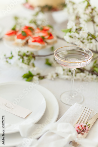 Table setting for a dinner with flowering cherry tree branches on the table. Vintage champagne glasses on the white linen tablecloth