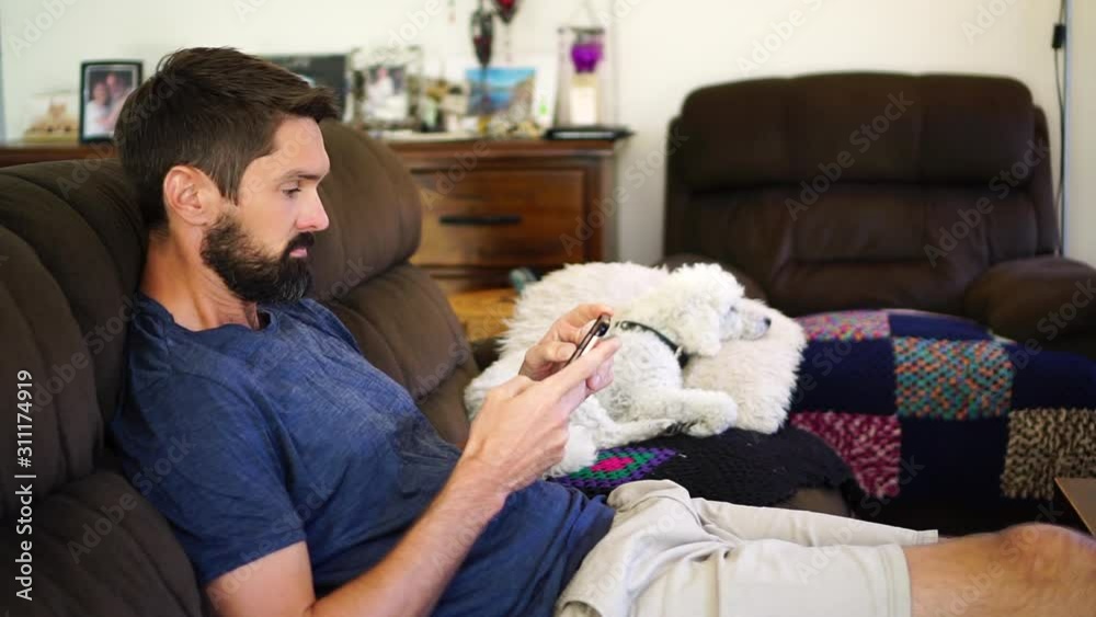 Caucasian man relaxing on sofa using smart phone, dog in background, medium shot Caucasian man relaxing on sofa using smart phone, dog in background, medium shot