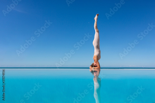 Charming young woman standing in salamba sirsasana exercise, headstand pose, on edge of pool against blue sky. The concept of improving blood circulation in the head. Copyspace
