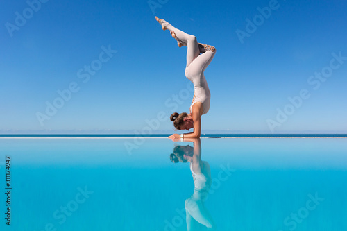 Charming young woman in a suit doing shirshasana standing on the edge of the pool against a blue sky. The concept of improving blood circulation in the head. Copyspace