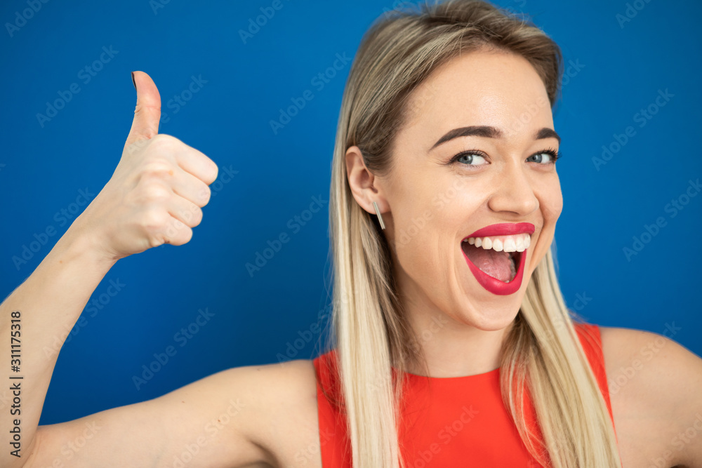 Happy emotional thumbs up beautiful blonde woman. Big adorable teeth smile. Red lips. Blue background Happy emotional thumbs up beautiful blonde woman. Big adorable teeth smile. Red lips. Blue background