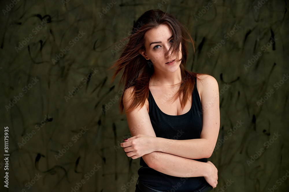 Fototapeta premium Beauty concept, stylish portrait. Photo of a pretty glamorous brunette girl in a black t-shirt with flying hair on a gray fashionable original background.