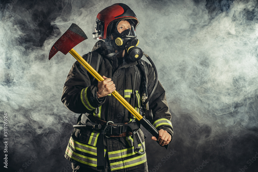 Obraz premium portrait of serious and confident caucasian fireman stand holding hammer, wearing special protective uniform in the smoky background