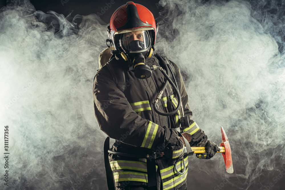 Obraz premium portrait of serious and confident caucasian fireman stand holding hammer, wearing special protective uniform in the smoky background