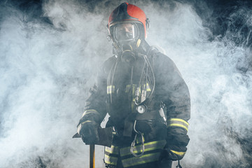 young fireman in protective coat using special equipment for fire fighting, isolated over smoky background