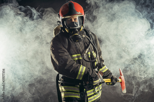portrait of serious and confident caucasian fireman stand holding hammer, wearing special protective uniform in the smoky background