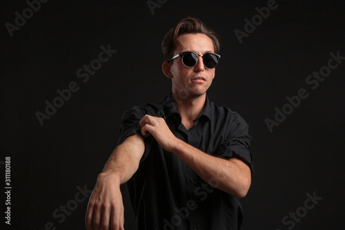 Confident stylish man in black eyeglasses buttoning up balck shirt isolated over black background