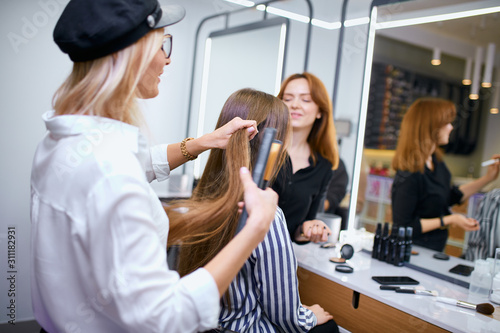 professional hair stylist making hairdo to young caucasian girl with long hair, careful hairdresser and young beautiful client in beauty saloon